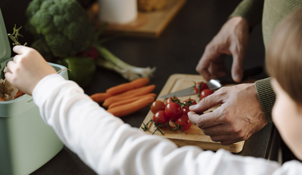 Auf dem Bild ist im Vordergrund ein Mädchen mit einem weißen Pullover zu sehen, dass etwas in einen hellgrünen Kübel entsorgt. Im Hintergrund schneidet eine Männerhand Tomaten.